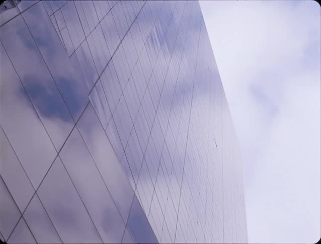 A view looking up from the street shows white clouds and tall buildings reflected in the dark glass windows of a modern skyscraper under a blue sky