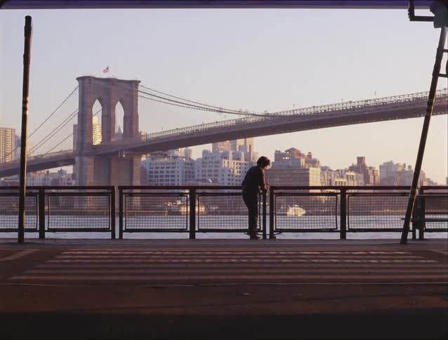 Young adult man standing on a lookout over a body of water next to a bridge and a metropolitan area then skating away outdoors during dusk or dawn
