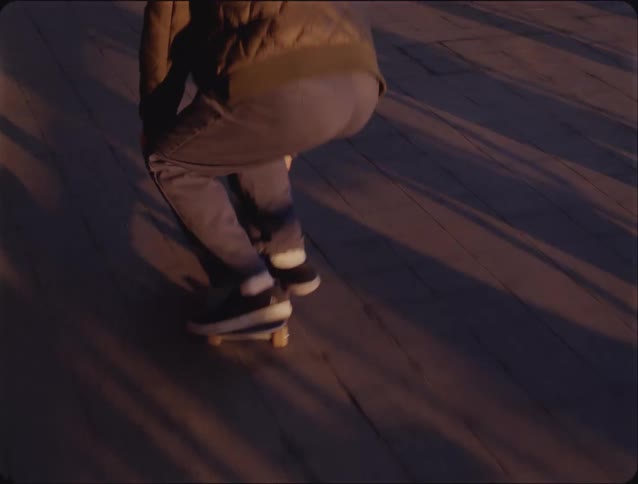 A young man riding a skateboard along a waterfront path beneath an overhead bridge at sunset