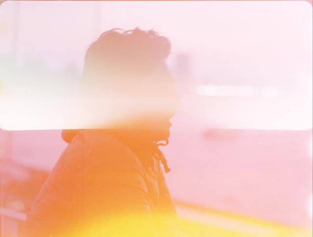 A close view shows a man with dark hair and a jacket leaning on a railing while looking out at a city harbor and river during a purple sunset