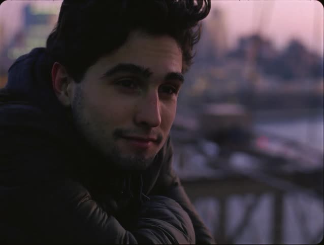 A close view shows a man with dark hair and a jacket leaning on a railing while looking out at a city harbor and bridges during a purple sunset