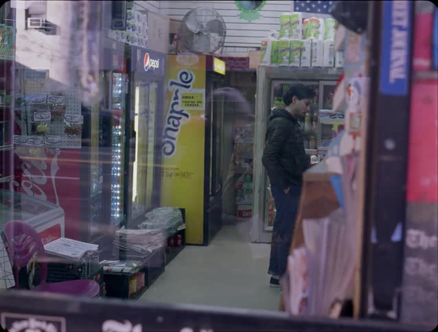 A man wearing a dark jacket and blue pants stands inside a small convenience store and looks through products near a yellow drink cooler and glass display cases while cars pass by outside