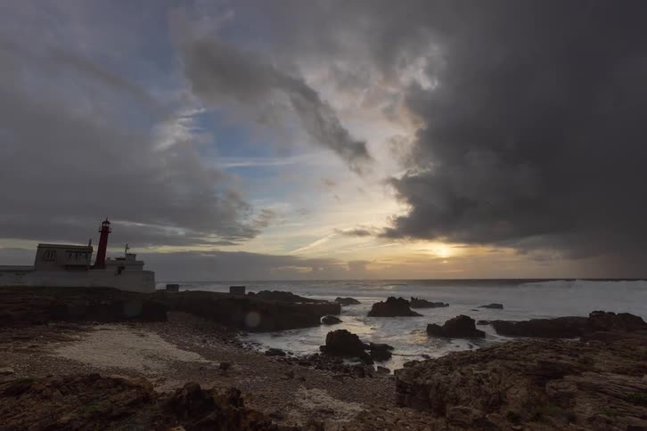 Red lighthouse beside white building on rocky coast with sea and clouds at sunset