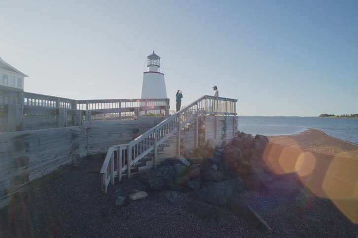 Two tourists stand on wooden stairs near a lighthouse overlooking the ocean on a sunny coastal day