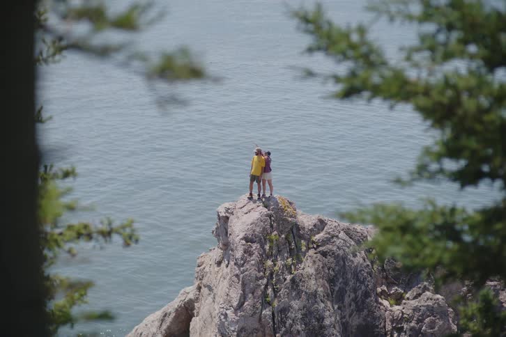 Two people standing high on a rock formation along the coast overlooking the water