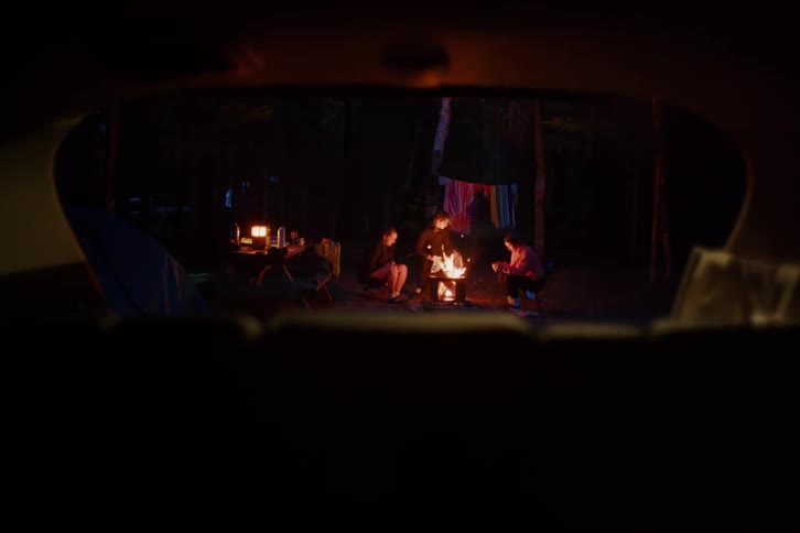 A group of friends sits around a glowing campfire at night in a dark forest as seen through the open back of a car