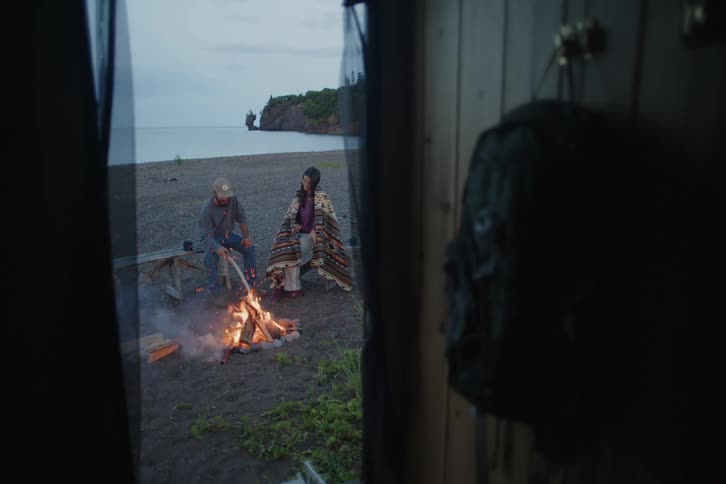 A man and woman sit by a glowing campfire on a dark sand beach at dusk viewed through a wooden cabin doorway