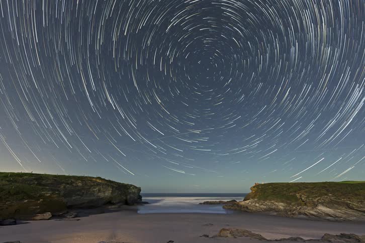 A time lapse shows stars rotating in a circular pattern over a sandy beach and dark ocean waves between rocky cliffs during a clear night