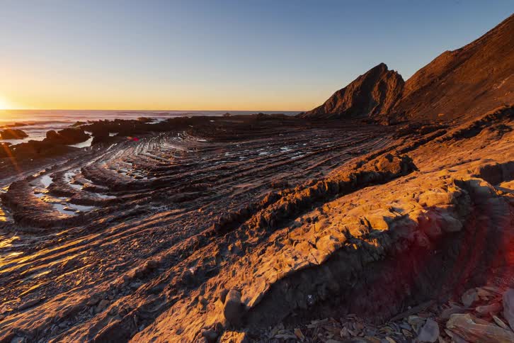 Golden sunlight stretches across rugged coastal rock formations as waves roll in beside layered cliffs under a clear evening sky