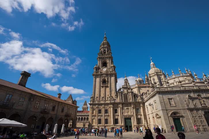 Tourists gather in a large plaza in front of a historic cathedral with a tall bell tower under a bright blue sky