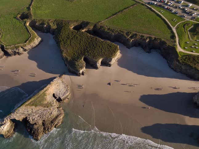 Ocean waves reaching a beach between rocky cliffs with fields and houses in the background