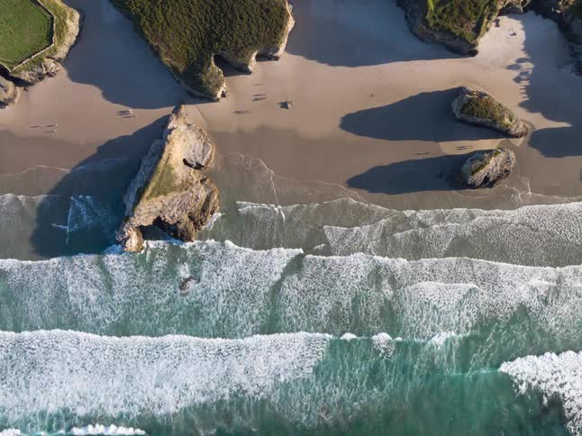 A beach with waves crashing on the shoreline people walking along the sand and a large rock formation sitting on the shore