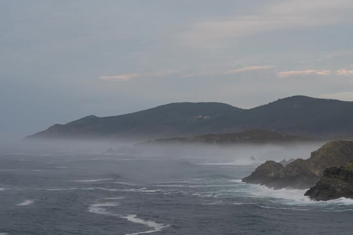 Ocean waves crash along a rocky coastline with misty mountains in the distance under an overcast sky