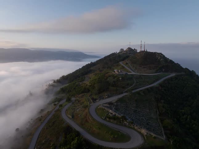 Winding road climbing a hill with communication towers at the top above a layer of clouds