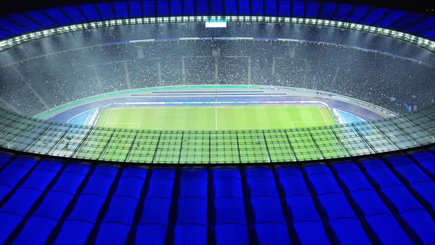 A massive soccer stadium at night with bright floodlights illuminating the green pitch and a full crowd of fans while a blue circular roof frames the brightly lit field during a professional match