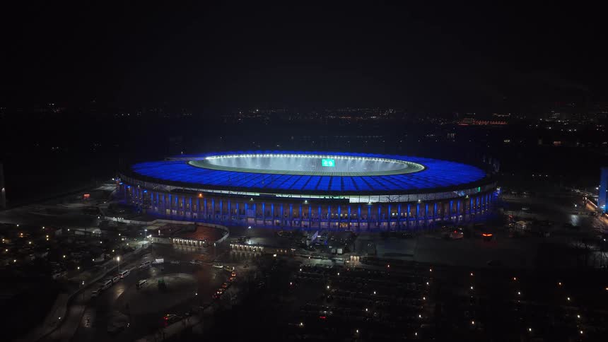 Brightly lit stadium with a blue roof sits in a city at night
