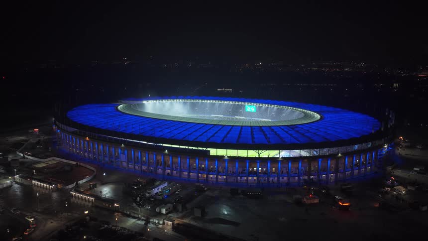 Circular stadium illuminated in blue at night with stands and field visible