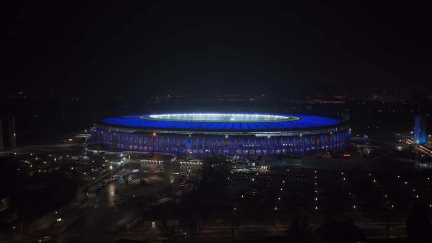 A wide shot of a large circular stadium illuminated with blue lights at night
