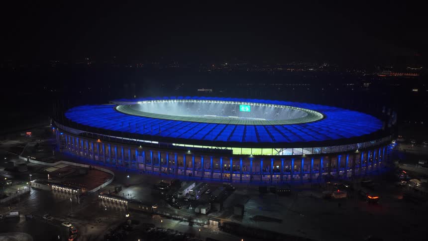 An aerial view shows a large stadium at night featuring a distinctive oval roof illuminated with bright blue lights bright white spotlights shine upward from the center of the field while the surrounding city remains dark