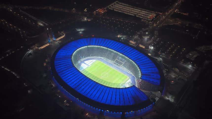 Illuminated oval stadium at night with a soccer field and surrounding city streets