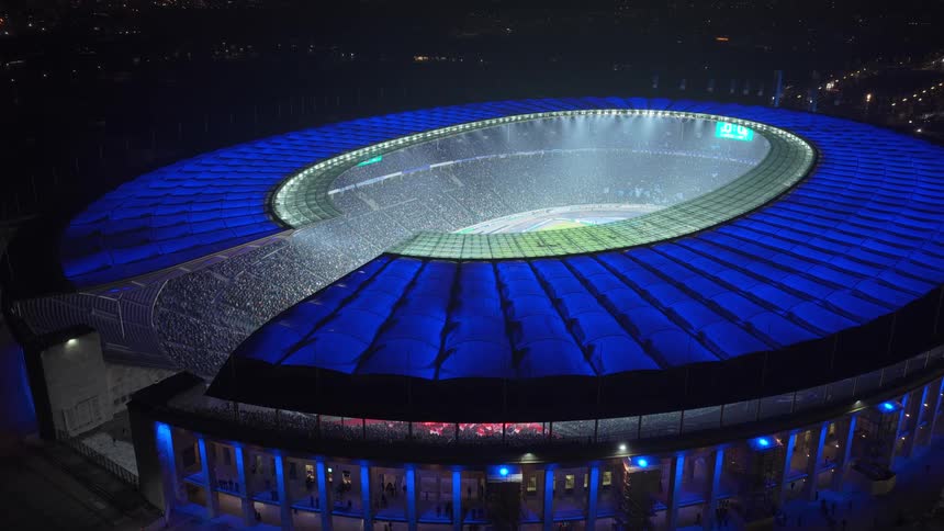 A brightly illuminated modern stadium glows blue at night packed with spectators and viewed from above in a dramatic aerial perspective