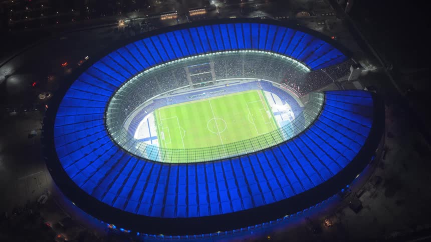 The video shows an aerial view of a football stadium at night featuring a glowing blue roof that circles the brightly lit green pitch the camera slowly zooms in on the field where players are positioned during a match