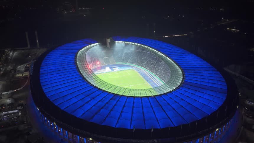 Aerial view of a brightly lit sports stadium at night with a glowing blue roof and illuminated field