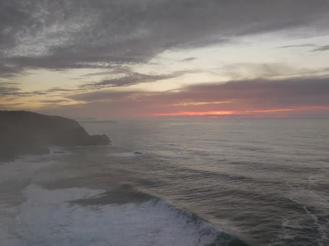 A drone flies over dark ocean waves breaking against a steep coastal cliff side while the sun sets on the horizon under a cloudy grey and orange sky