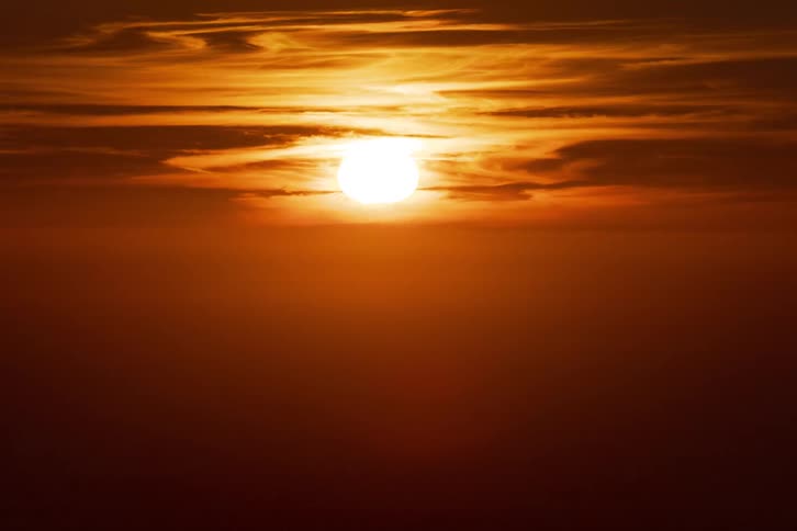 A close view shows a bright orange sun setting behind thin layers of clouds over a dark ocean while the sky glows with a warm golden light