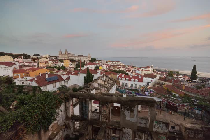 People and vehicles move through a coastal town in the area near a ruined building as night falls