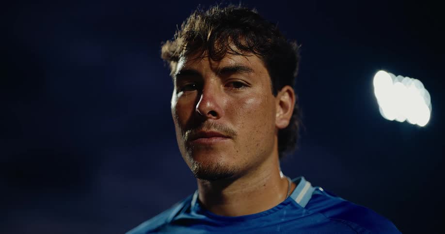 A man with dark wavy hair and a slight mustache looks directly into the camera with a steady serious expressionhe is positioned against a dark night sky illuminated by a bright stadium light wearing a blue athletic jersey