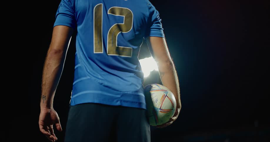 Adult male soccer player standing on a stadium holding a footbal under his arm outdoors during night time