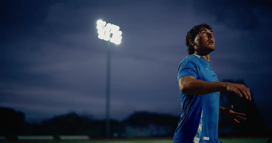 A soccer player wearing a blue argentina jersey with the number 12 practices under stadium lights at dusk he focuses intently on the ball looking upward before executing a kick as the camera captures his movement from a low angle