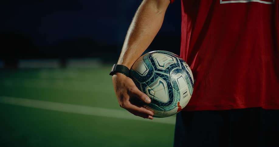 A male soccer player stands on a turf field at night holding a white and blue ball against his side while wearing a red jersey and a black watch on his wrist