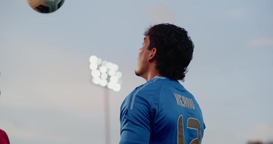 A close view shows a man in a blue sports jersey throwing a soccer ball into the air toward another man in a red shirt on a field under a pale sky