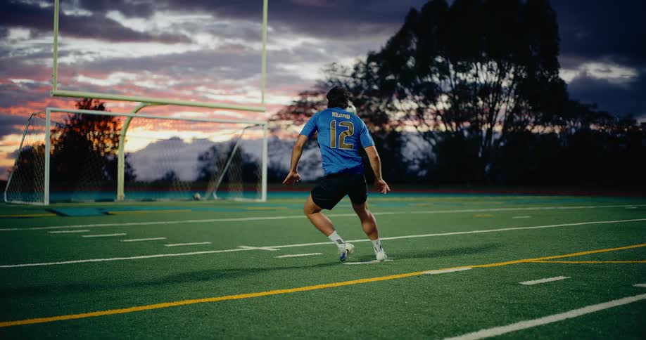 A soccer player wearing a blue number twelve jersey kicks a ball toward a goal on an artificial turf field during a cloudy sunset
