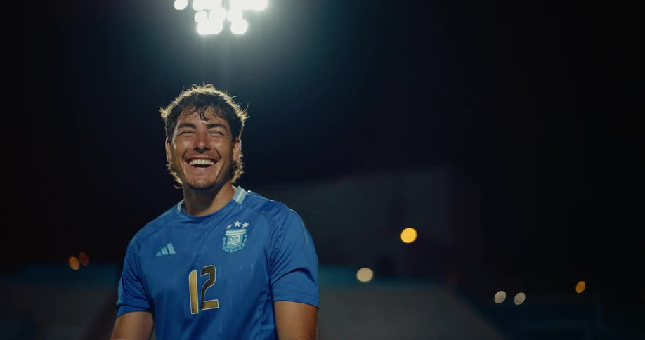 A smiling male soccer player wears a blue argentina jersey with the number twelve while standing on a stadium pitch at night under bright floodlights as he holds a ball
