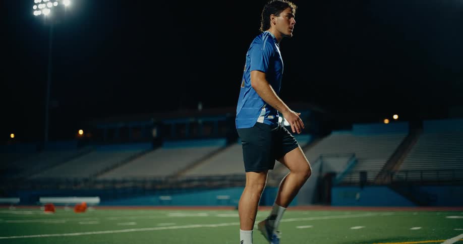 A man wearing a blue sports shirt and black shorts kicks a soccer ball on a green field at night under bright stadium lights and then walks away