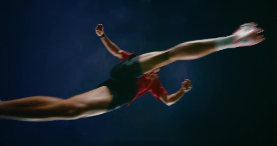 A view from below shows a man in a red sports jersey jumping and performing a bicycle kick against a dark background during a soccer match