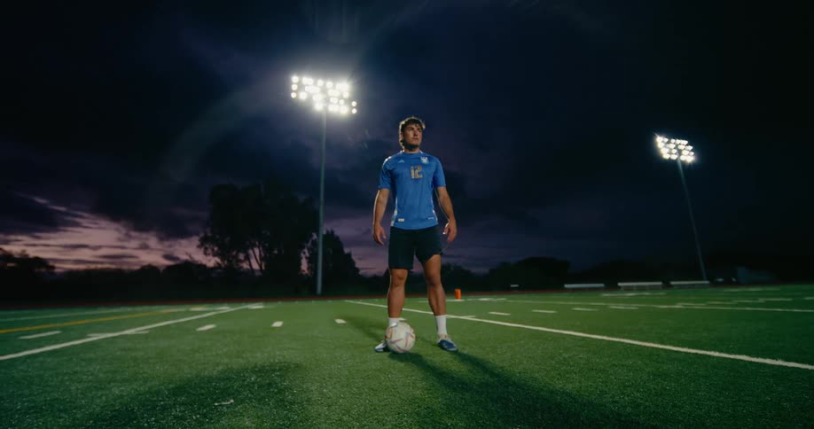 A soccer player stands with a ball on a lit outdoor field at night under stadium lights