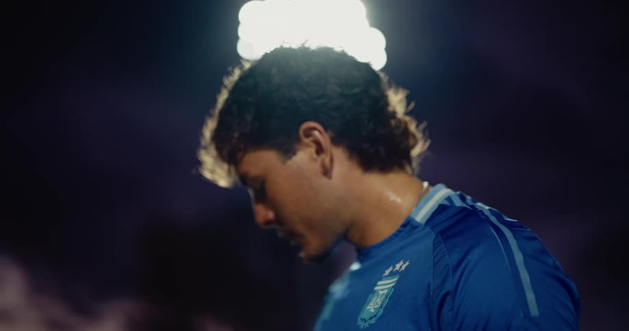 A athlete in a blue jersey is shown in side profile under bright stadium lights with sweat visible on his face during the match