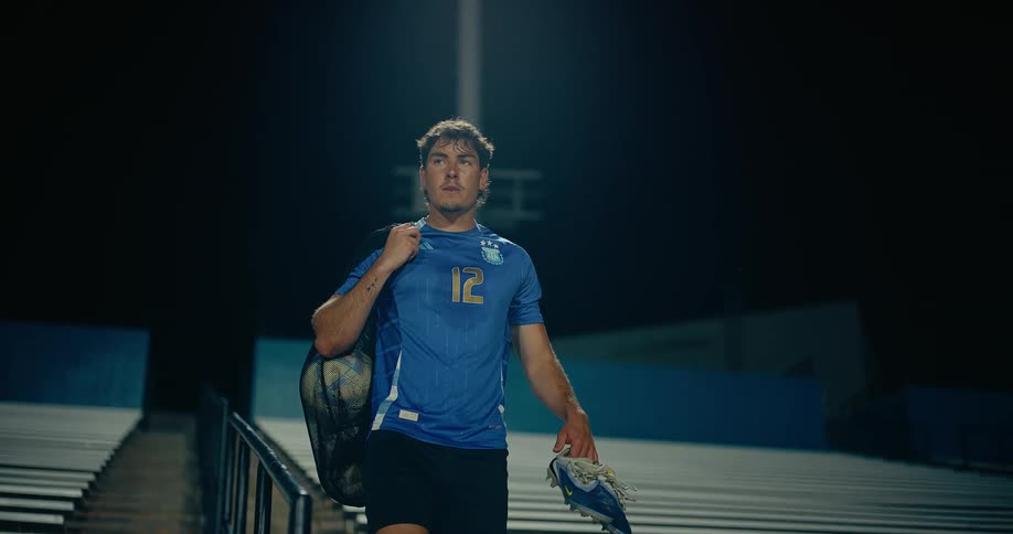 Young man walks down stairs carrying his shoes with a bag of soccer balls over his shoulder