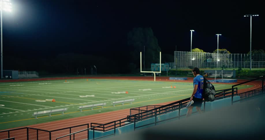 Young man walks down stairs toward a sports game field carrying his shoes with a bag of soccer balls over his shoulder