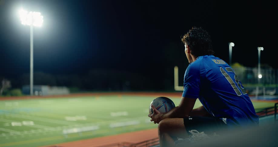 Player wearing blue number 12 jersey sitting while holding a ball on a sports field illuminated at night
