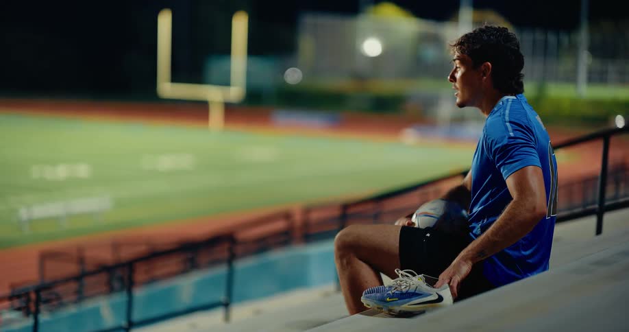 Male soccer player sitting in the stands of a football field stands while holding a shoe and a soccer ball