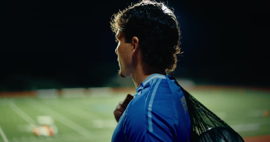 A focused soccer player stands under bright stadium lights at night catching his breath with a training bag over his shoulder as the quiet field stretches behind him