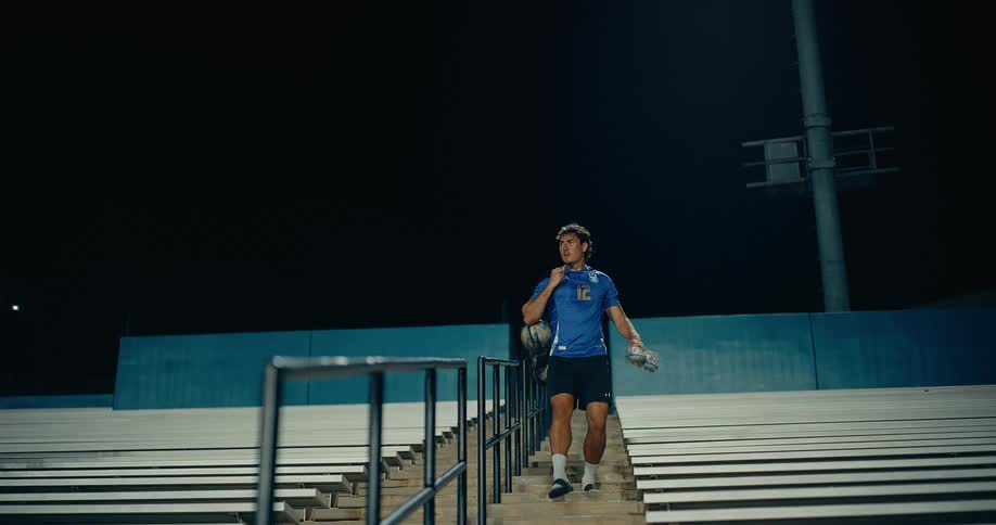 Young man walks down stairs carrying a pair of shoes with a bag of soccer balls over his shoulder