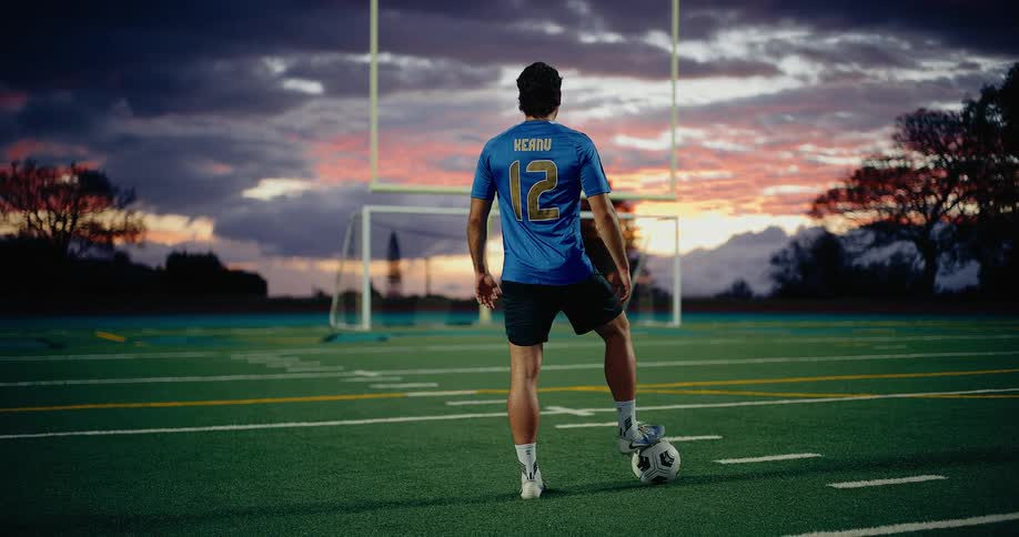 A lone soccer player wearing a blue number 12 jersey stands on the field at sunset with one foot on the ball facing the goal as dramatic clouds glow in the evening sky
