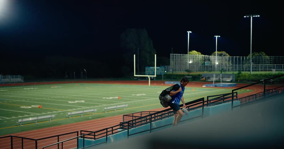 An athlete walking up the staircase inside a football stadium at night carrying a bag with a ball inside