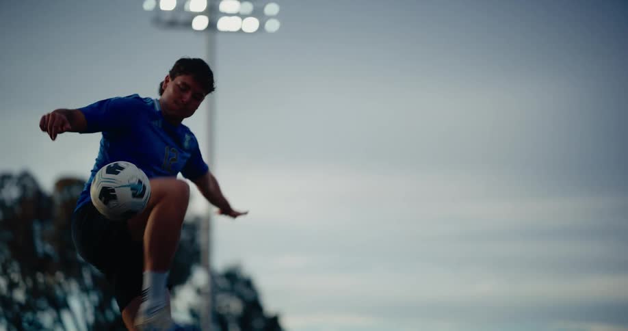 A focused soccer player in a blue shirt skillfully juggles the ball under bright stadium lights as evening settles over the field